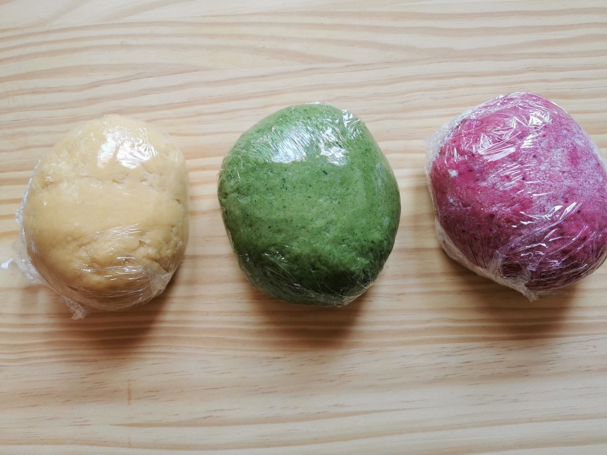 3 balls of colored pasta dough wrapped in plastic wrap on wood work surface.