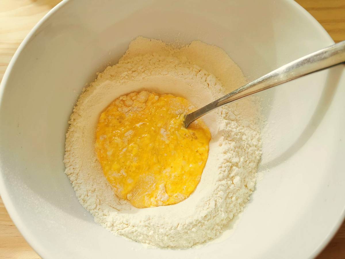 Beaten eggs in the middle of a mound of flour in white bowl.