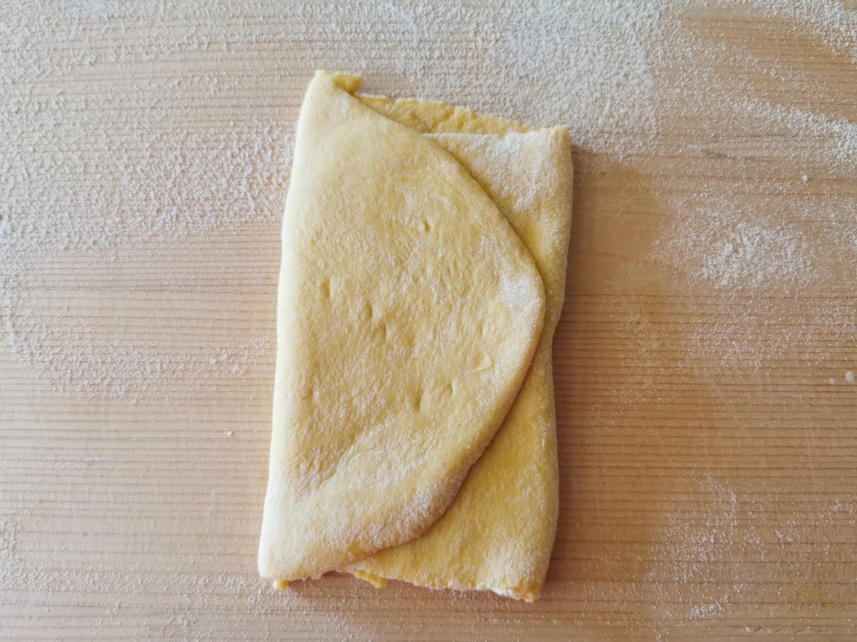 Piece of pasta dough folded into thirds on flour dusted wood work surface.