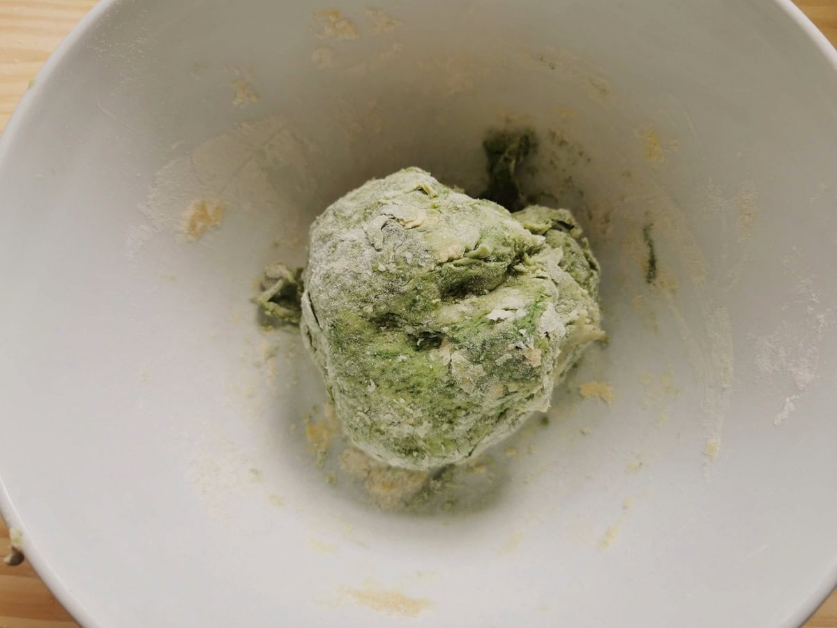 Rough ball of green pasta dough in white bowl.