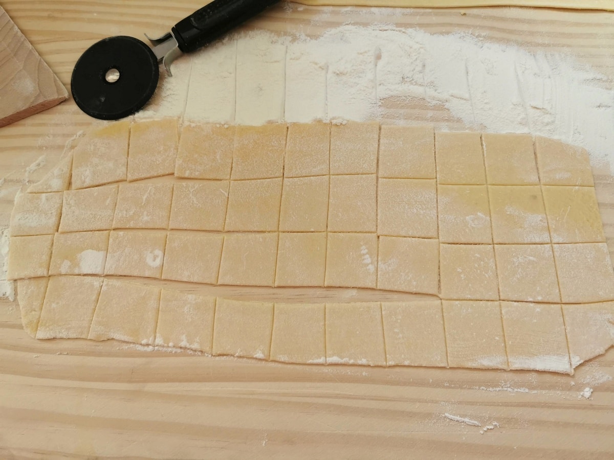 Fresh pasta dough sheet cut into small squares on wood pastry board.