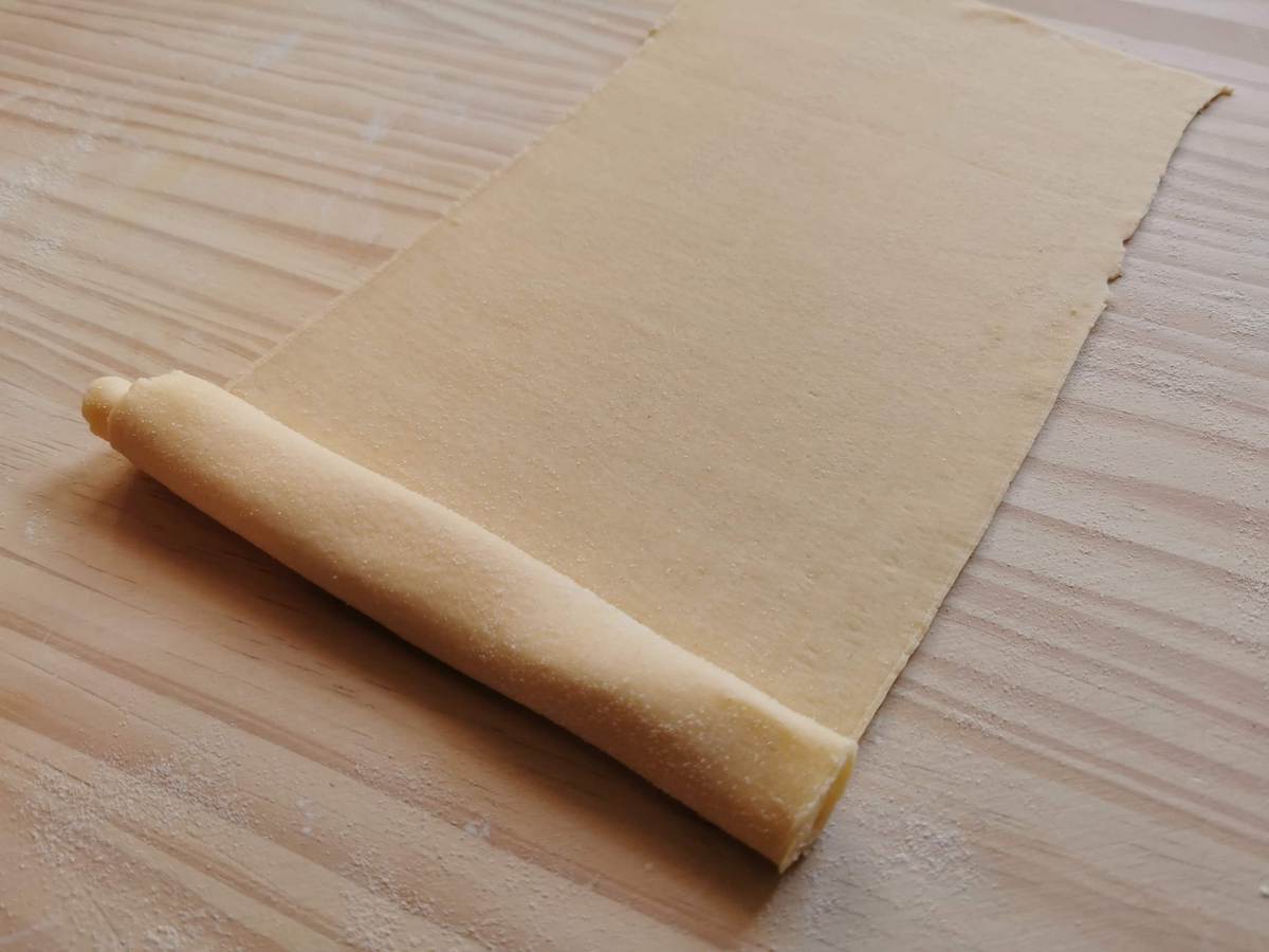 Rectangular pasta dough sheet half rolled up on wood worktop.