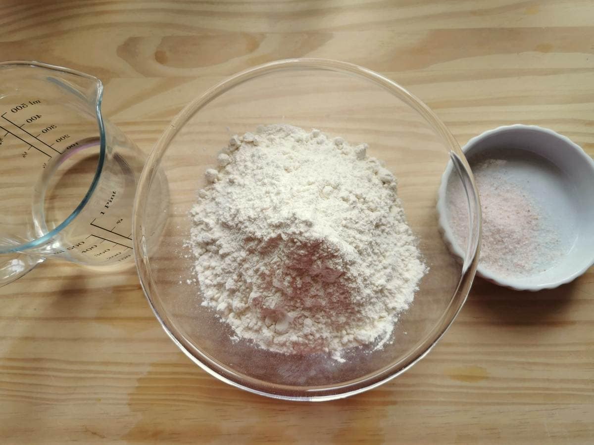 Ingredients for homemade strangozzi pasta on wood worktop;flour in a glass bowl, salt in a small white bowl and water in a jug.