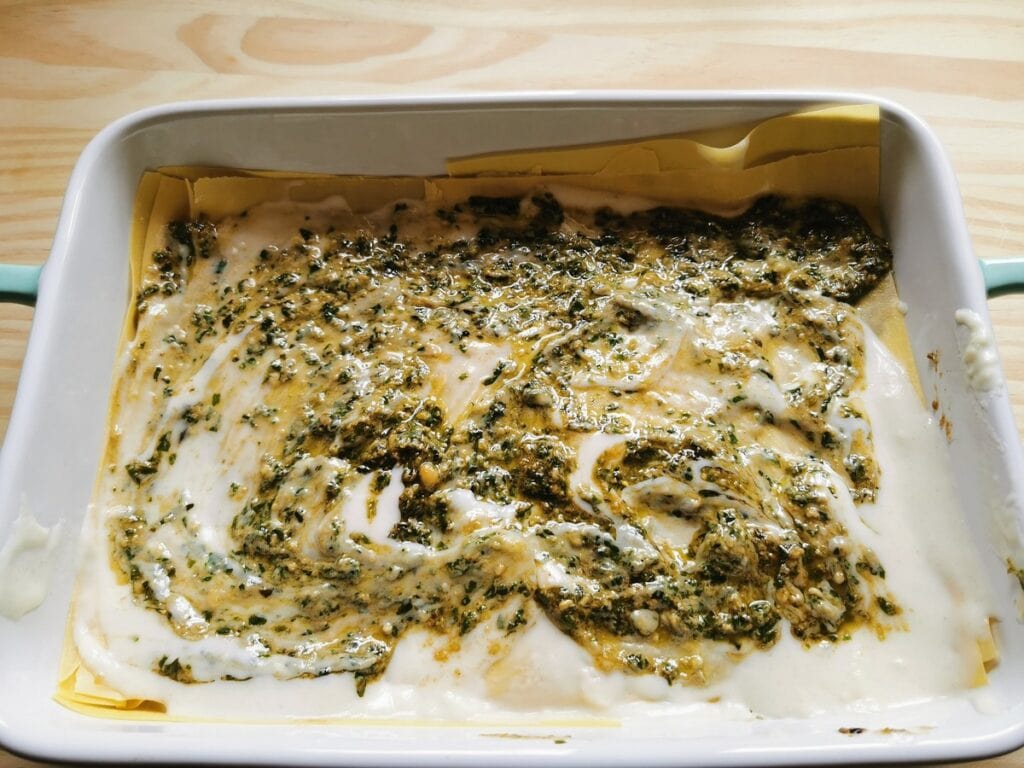 Béchamel and pesto spread over lasagna sheets in oven dish.