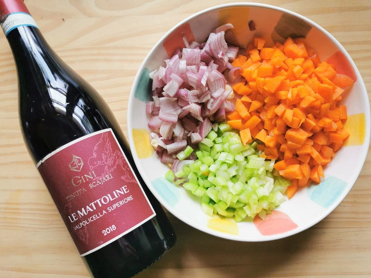 Peeled and cubed onion, celery and carrot in white bowl next to a bottle of red wine.