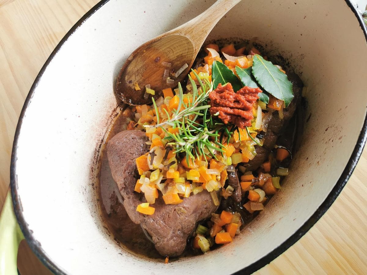 Beef cheeks with vegetables, tomato concentrate and herbs in Dutch oven.