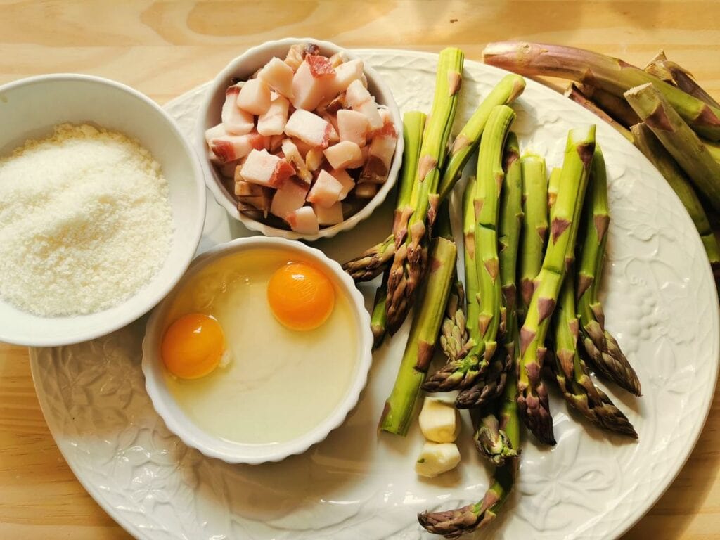 cubed guanciale in white bowl, grated pecorino in white bowl, 2 opened eggs in white bowl and cleaned asparagus spears on white plate