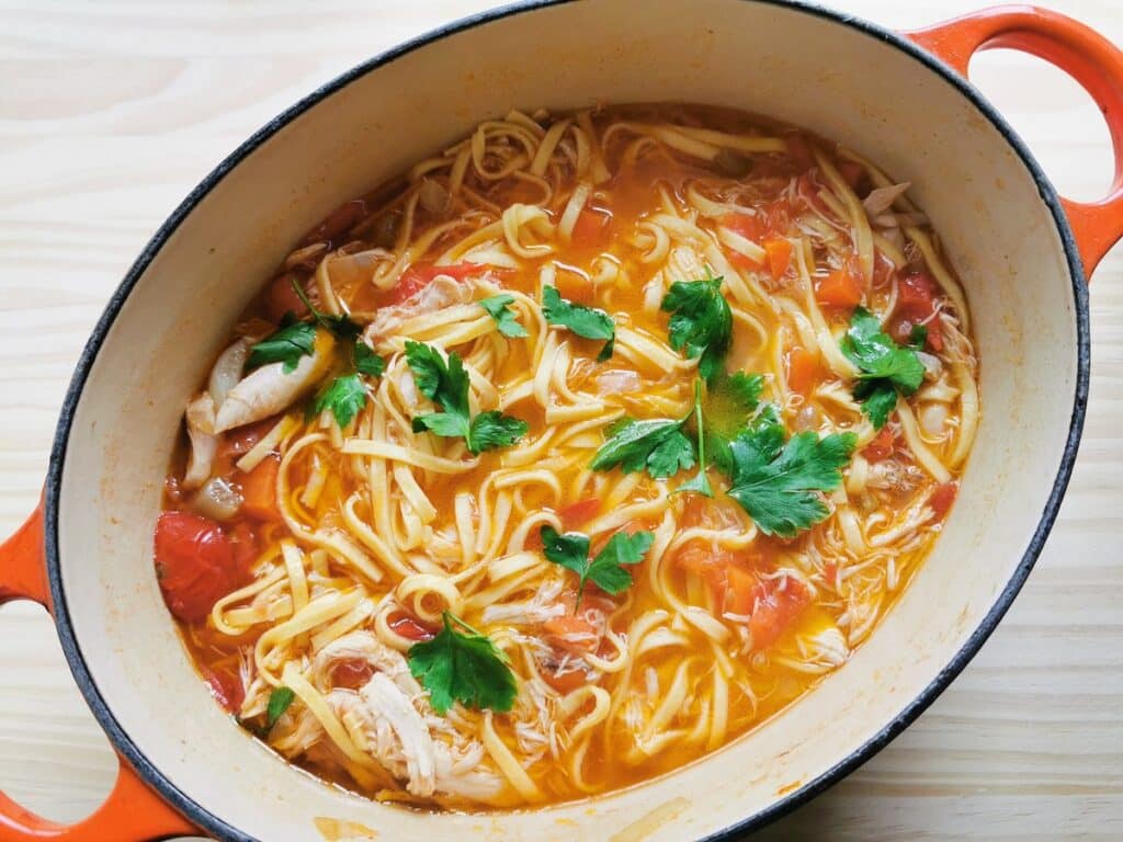 A large orange pot filled with noodle soup. The soup contains noodles, chunks of tomato, and pieces of chicken, garnished with fresh cilantro leaves. The pot is on a light wooden surface.