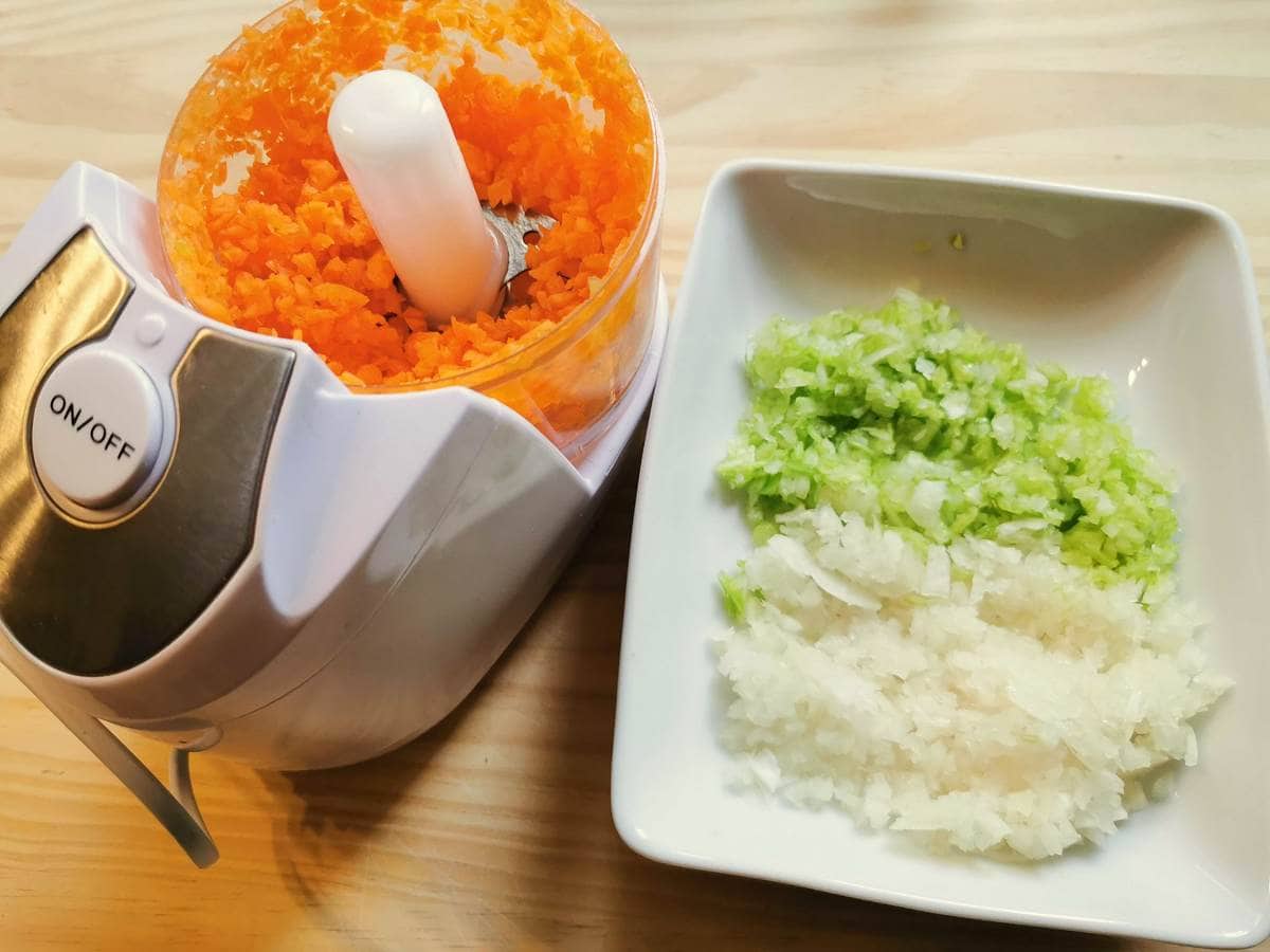 Finely chopped onion and celery in white bowl. And finely chopped carrots in food processor.