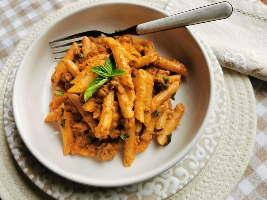 A bowl of penne pasta topped with a rich tomato and meat sauce, garnished with a fresh basil leaf. The dish is on a white plate with a white decorative edge, placed on a checkered tablecloth. A fork rests on the bowl's edge.