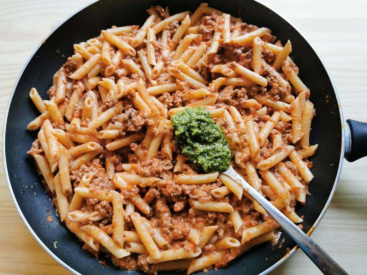 Tablespoon of pesto being added to pasta and ragu in frying pan.