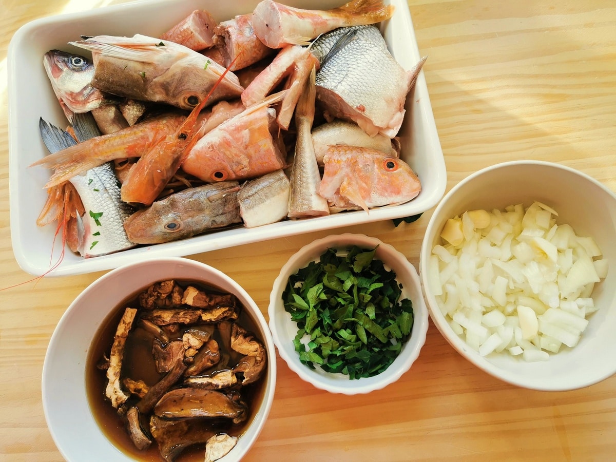 Prepared ingredients for this fish soup; cleaned and cut fish, chopped parsley, chopped onions and garlic, dried porcini in water in separate white bowls.