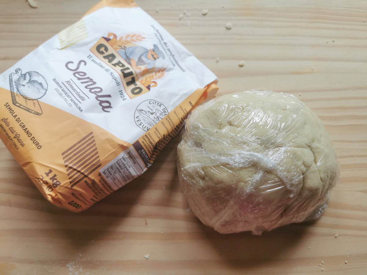 Ball of eggless pasta dough wrapped in plastic wrap on wood work top next to a bag of Italian semola rimacinata flour.