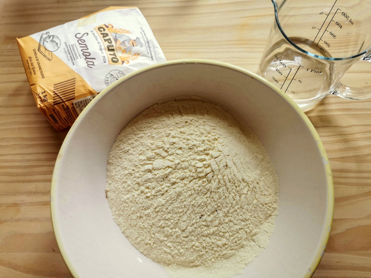 Hard wheat durum flour in large bowl with jug of water and a packet of semola rimacinata behind the bowl.