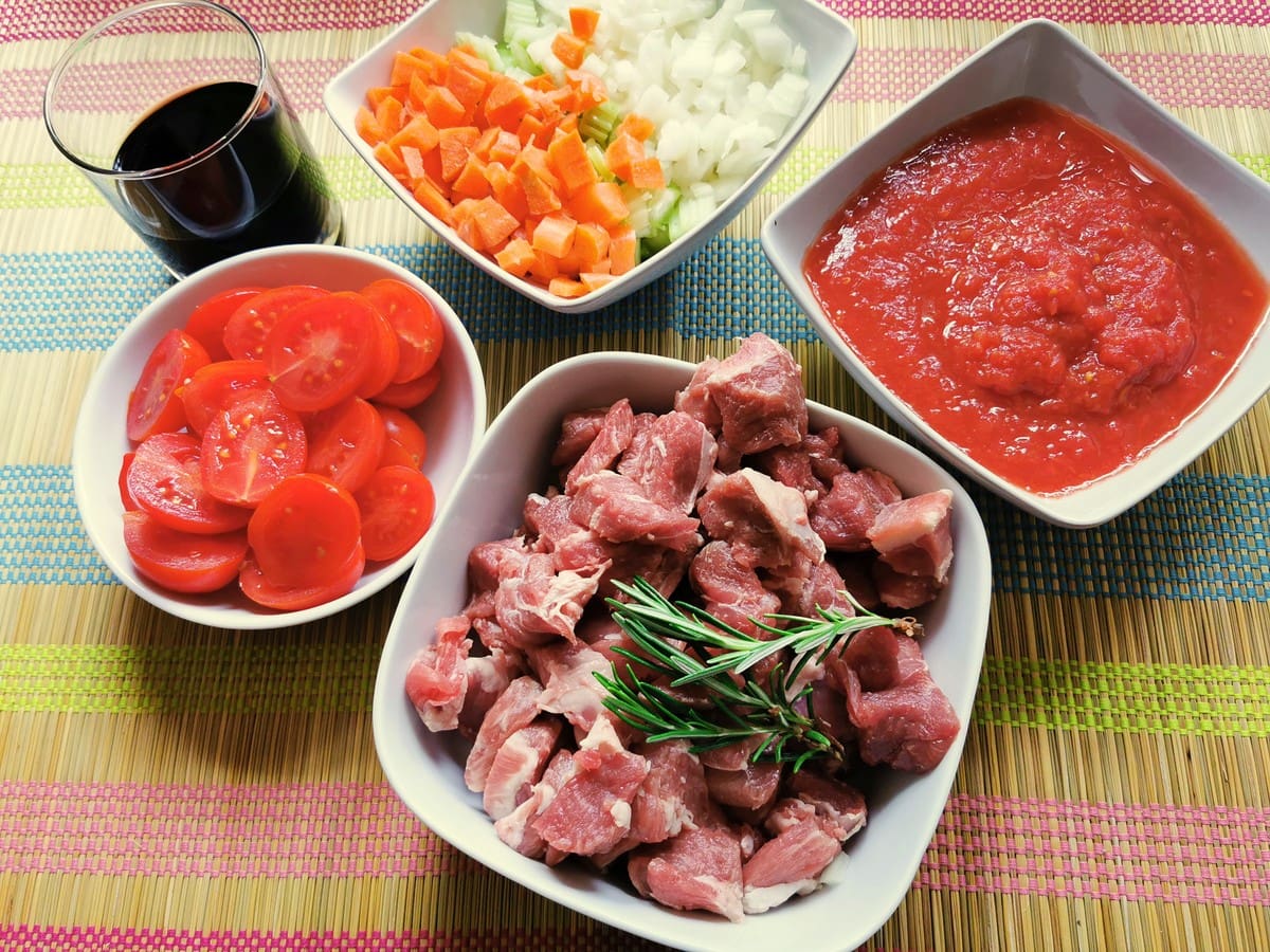 pieces of lamb, cut cherry tomatoes, chopped onions, clery and carrots and tomato passata all in separate white bowls