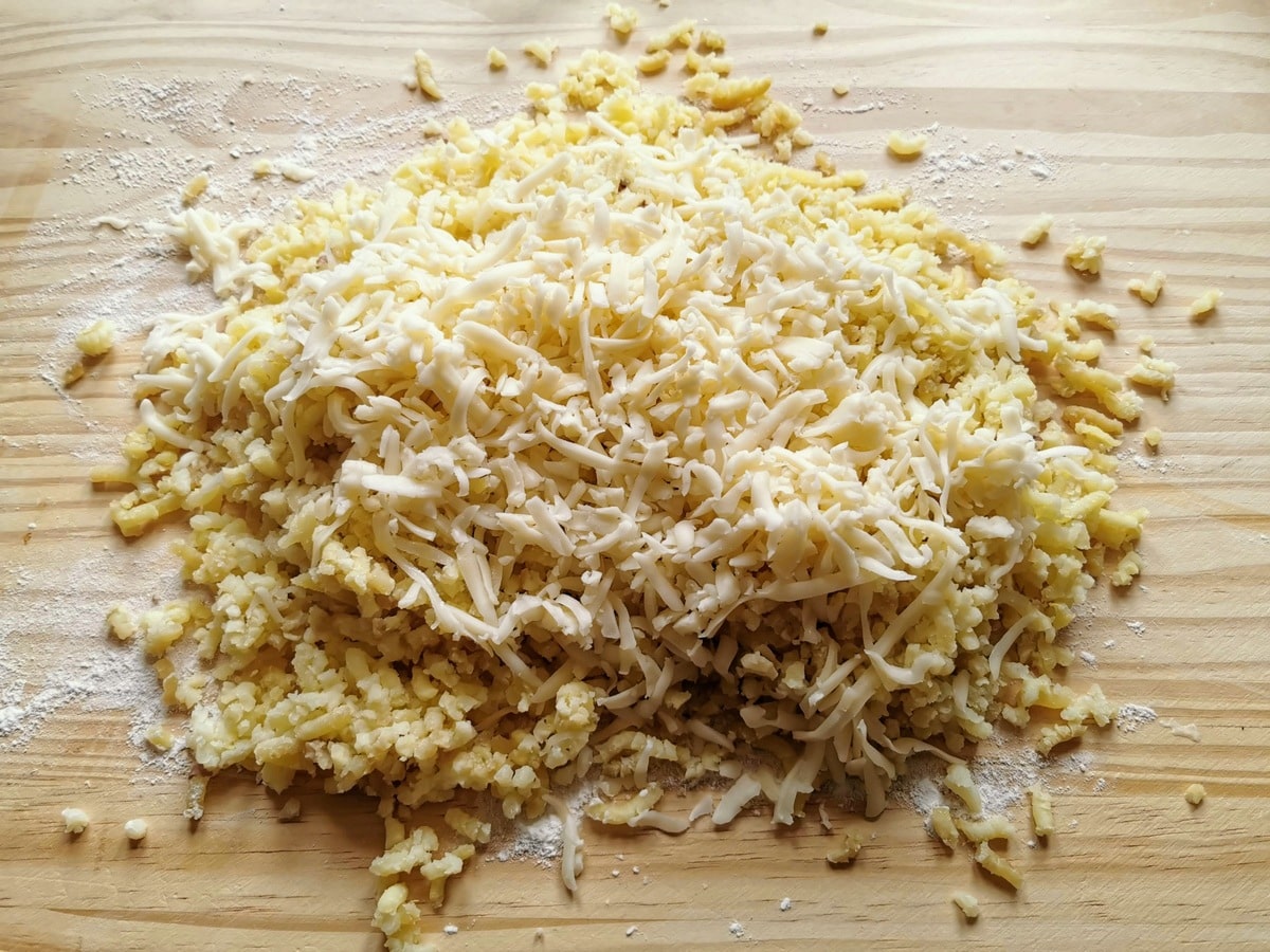 Grated Toma cheese added to riced potato on wood work surface.
