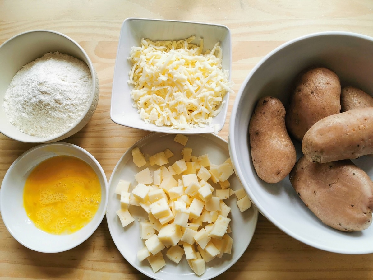 Boiled potatoes, grated cheese, cubed cheese, flour and a beaten egg all in separate white bowls.