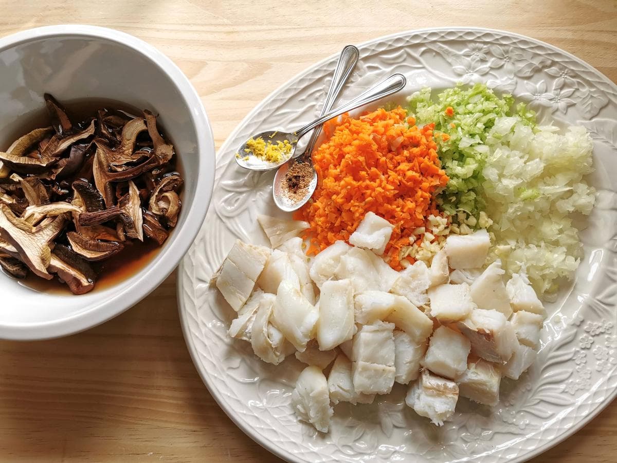 salt cod cut into chunks, finely chopped carrots, onion, celery and garlic on white plate and dried porcini soaking in water in a white bowl.