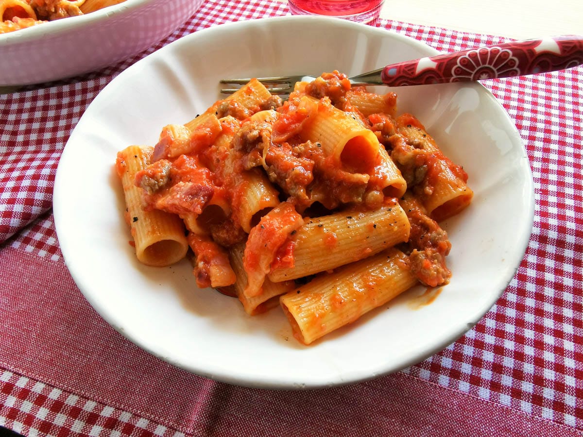 A portion of sausage rigatoni alla Zozzona in a white bowl on a checked red and white tablecloth.