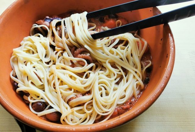 cooked linguine being mixed with baby octopus sauce in terracotta bowl