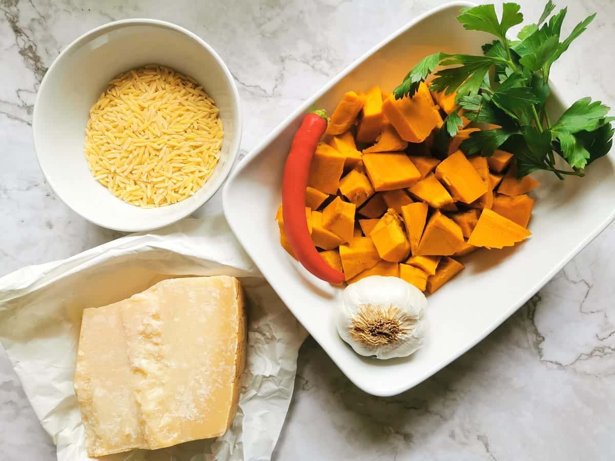 Ingredients for risotto style pasta with pumpkin. Cubes of pumpkin, parsley, red chilli pepper, garlic, risoi/orzo pasta and Parmigiano cheese on marble worktop.