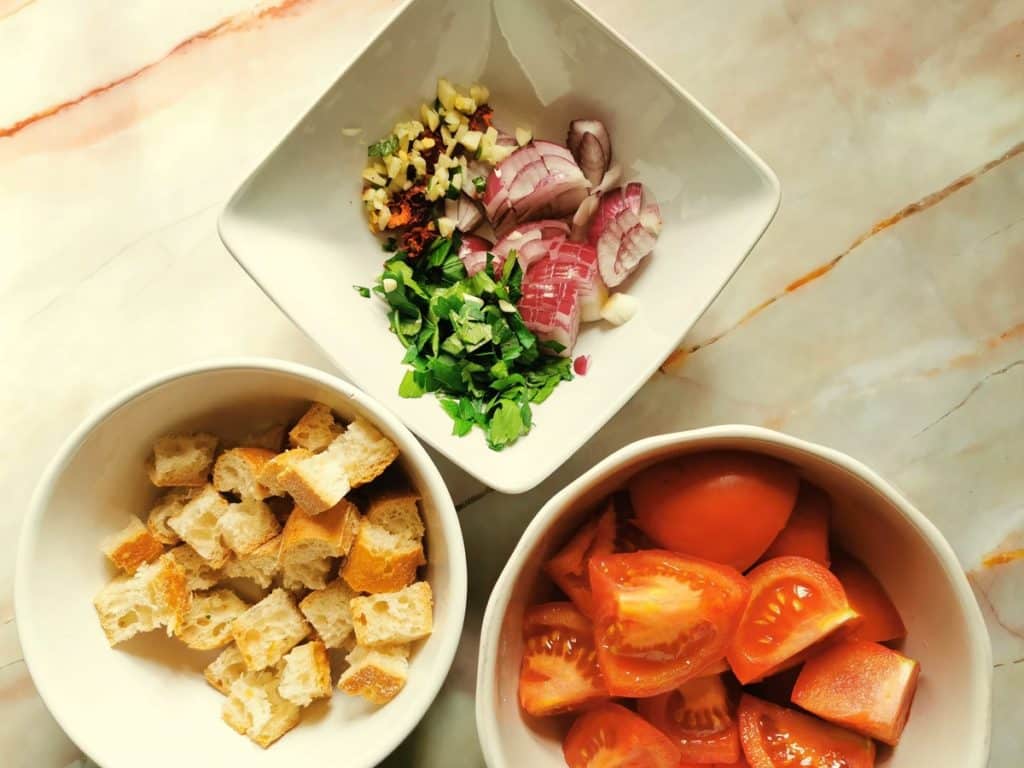 chopped tomatoes, cubed stale bread, chopped onions, garlic, parsley and chili pepper in white bowls.