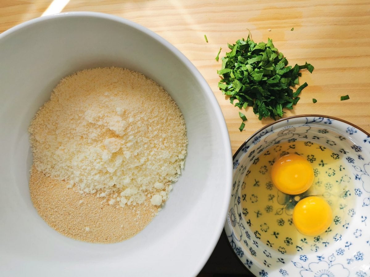 Breadcrumbs and grated cheese in large white bowl and 2 eggs opened into smaller bowl.