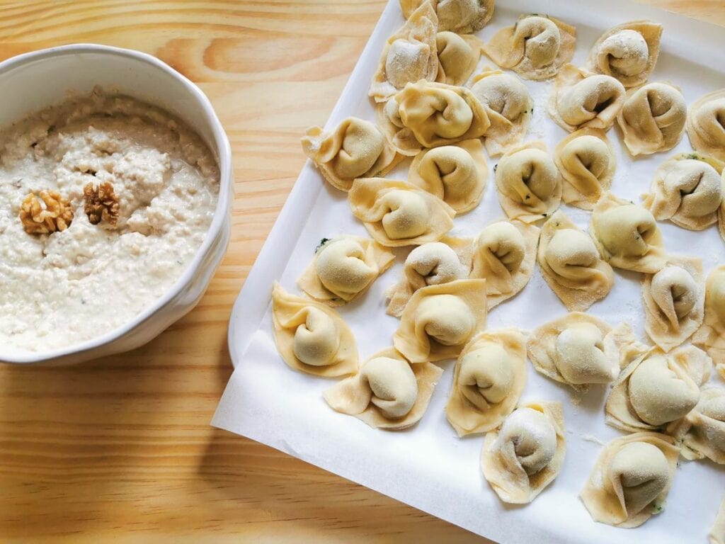 Ready walnut sauce in white bowl and uncooked pansotti on tray.