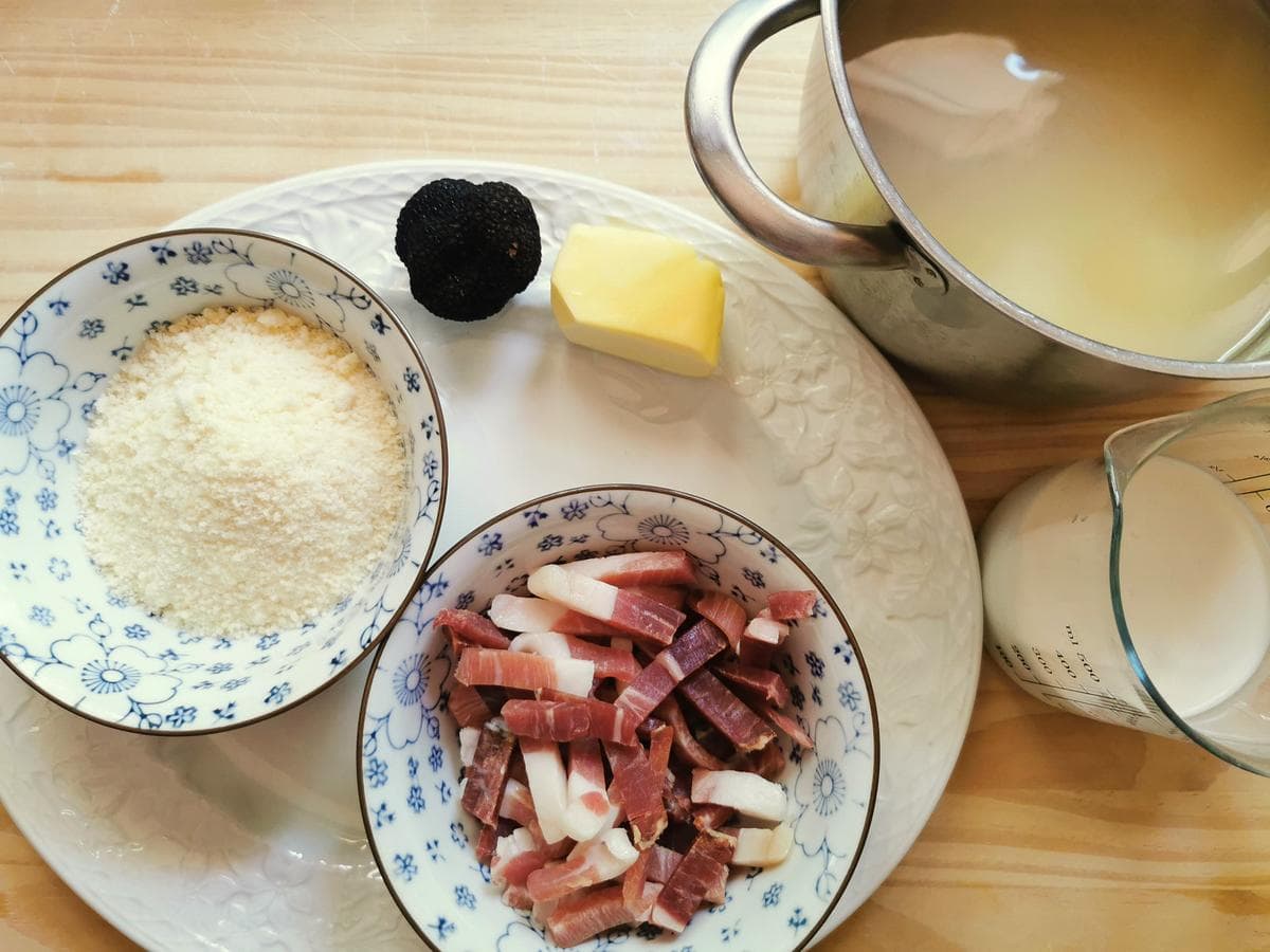 Strips of culatello in a bowl, grated Parmigiano in a bowl, broth in a pot and cream in a glass jug.