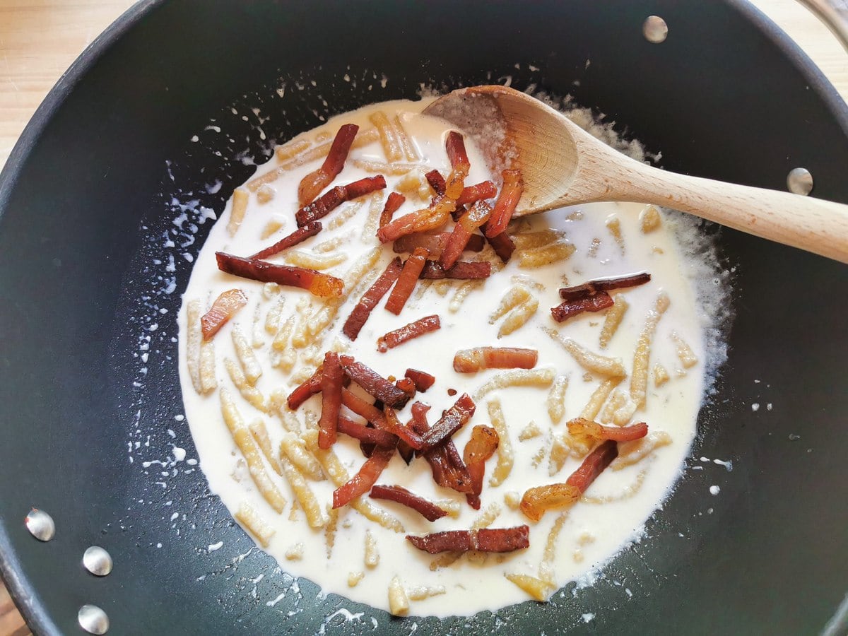 Fried prosciutto strips added to passatelli and Parmigiano cream sauce in deep skillet.