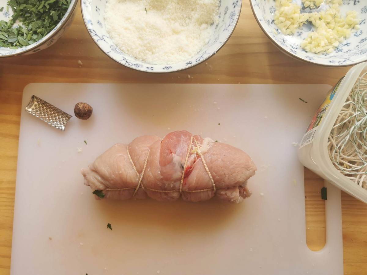One uncooked rolled pork involtini tied with kitchen string on white chopping board.