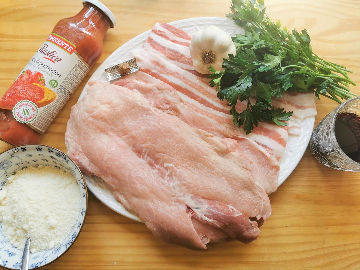 Ingredients for pork involtini on a white plate; pork slices, pancetta slices, garlic, fresh parsley, nutmeg, plus a glass of red wine and a jar of tomato passata.