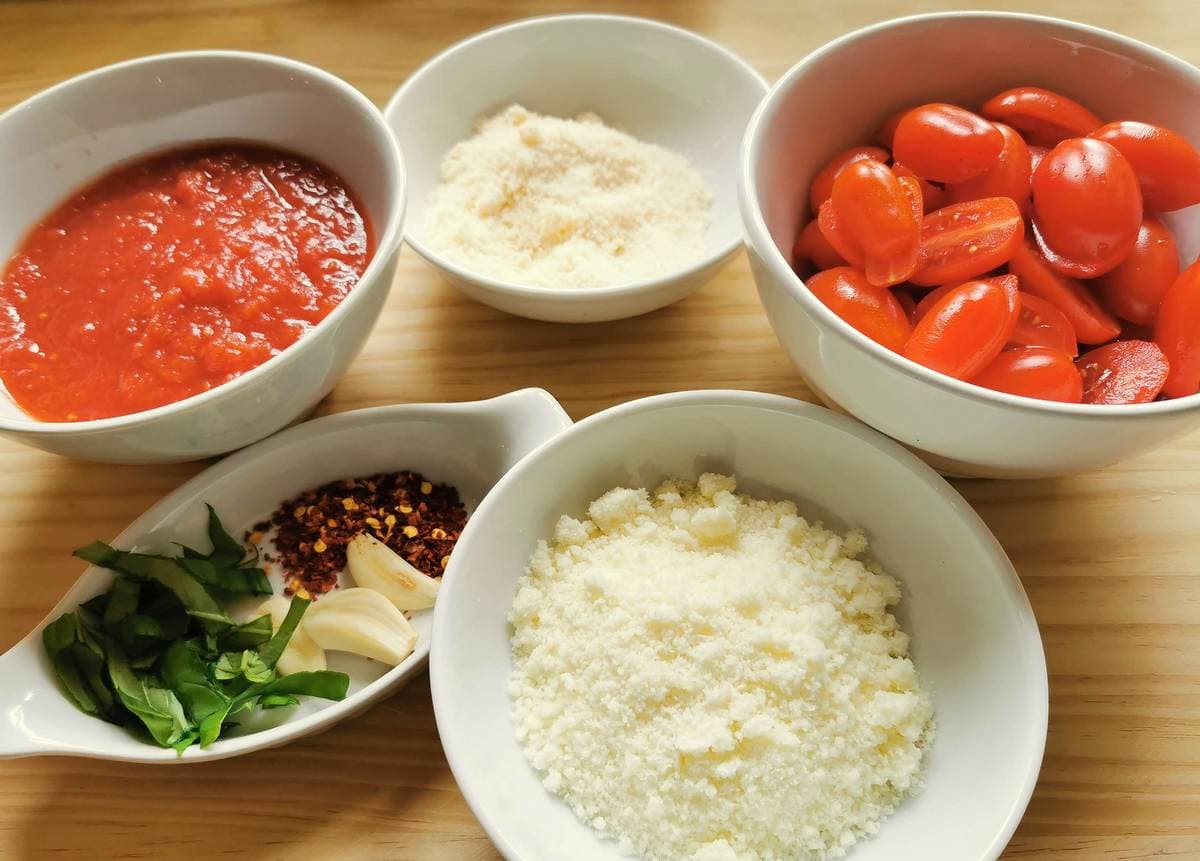 prepared ingredients, tomatoes, tomato passata, grated cheese, peeled garlic, chopped basil and peperocino flakes in separate white bowls on wooden work surface.