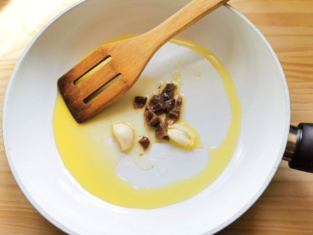 Garlic cloves and anchovy pieces cooking in olive oil in white frying pan