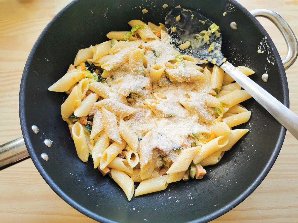 Grated grana added to the pasta and zucchini flower sauce in skillet.
