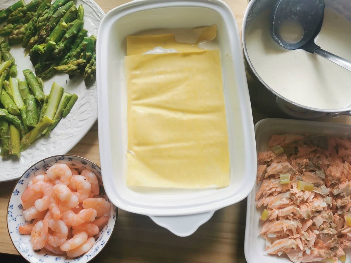 Fresh lasagne sheets in oven dish on top of layer of béchamel with shrimps, asparagus, and poached salmon in separte bowls.