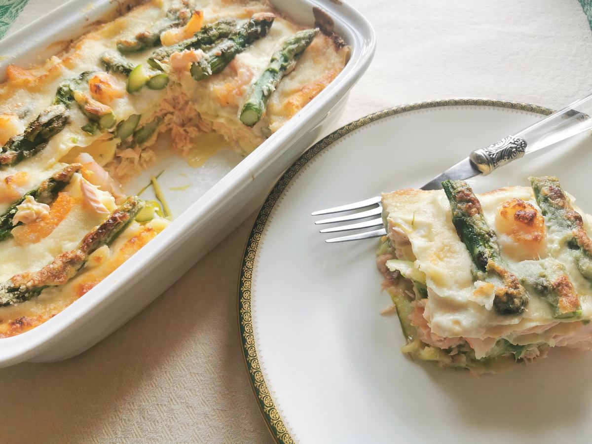 Baked poached salmon and asparagus lasagne al forno in oven dish with one portion ona white plate.
