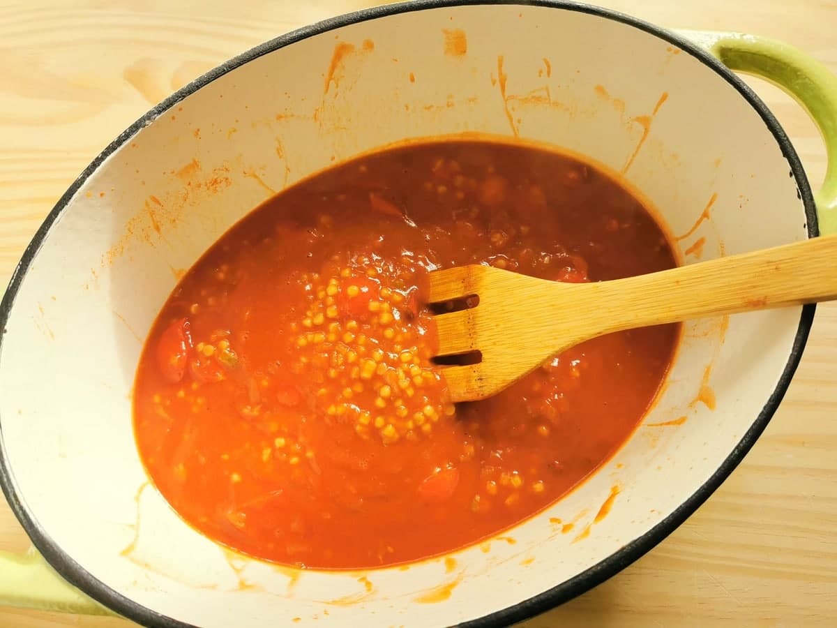 Fregola cooking in tomato sauce and broth in Dutch oven.