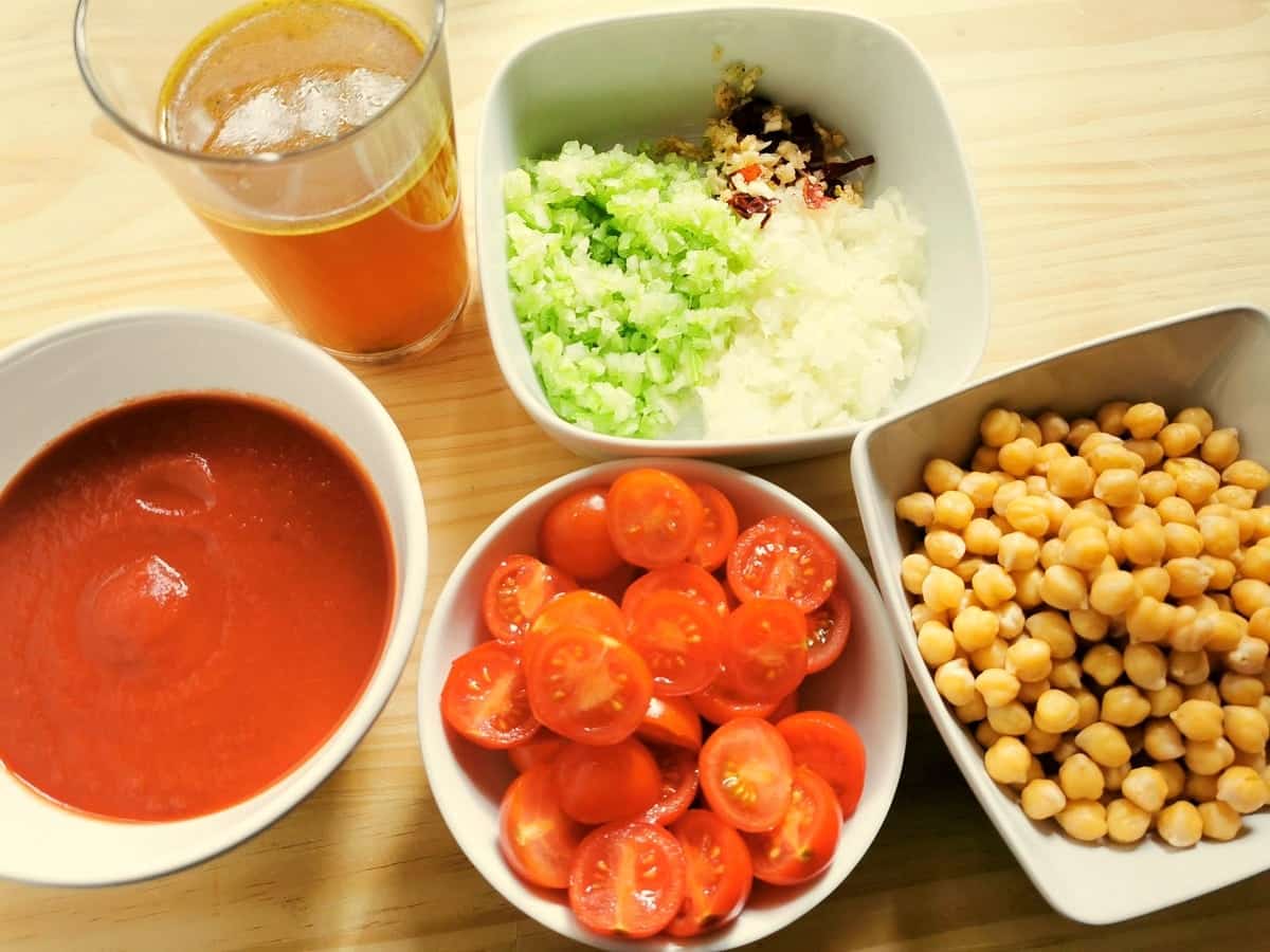 Celery, onion, garlic and peperoncino finely chopped in white bowl. bolied chickpeas in white bowl. Halved cherry tomatoes in white bowl and tomato passata in white bowl.