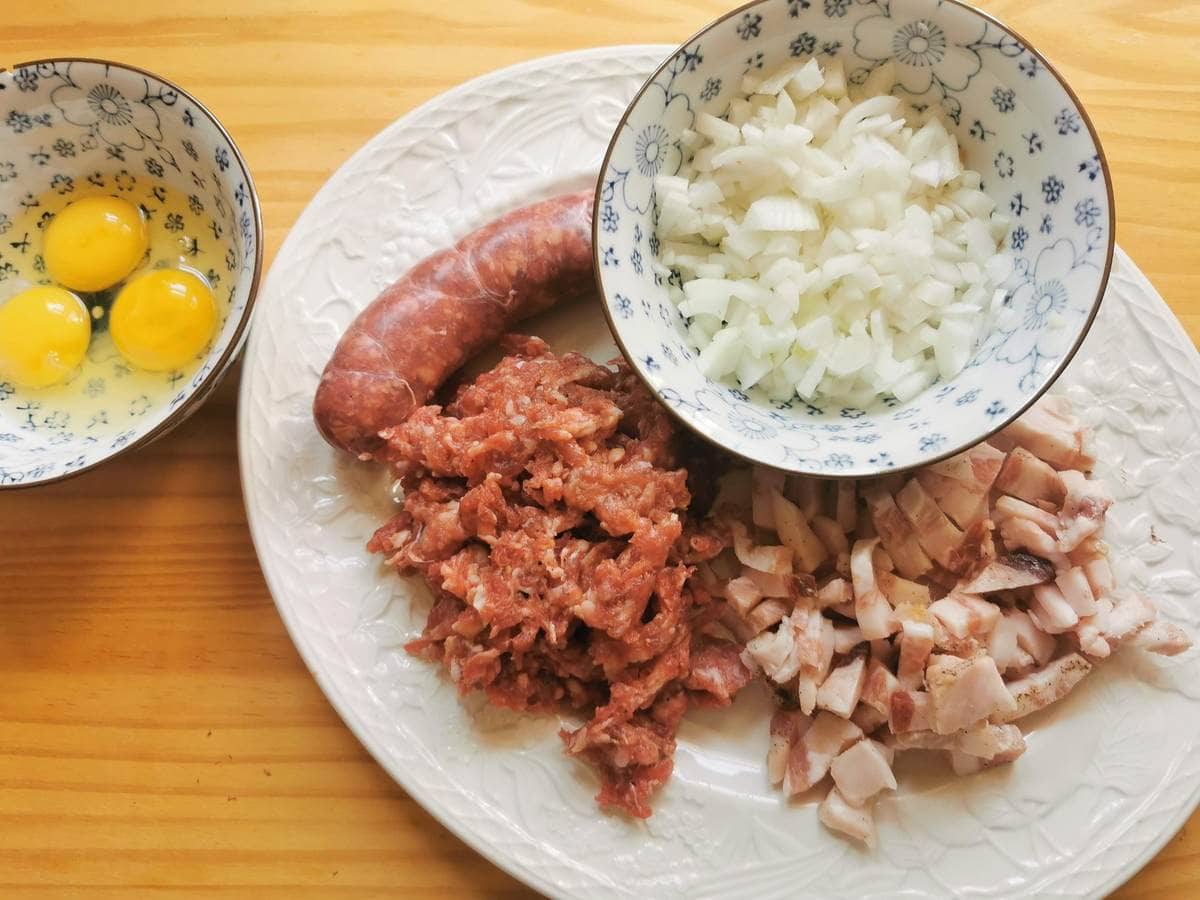 Peeled and sliced onion in small bowl, crumbled sausage meat and cubed guanciale on white plate and 3 egg yolks in small bowl.