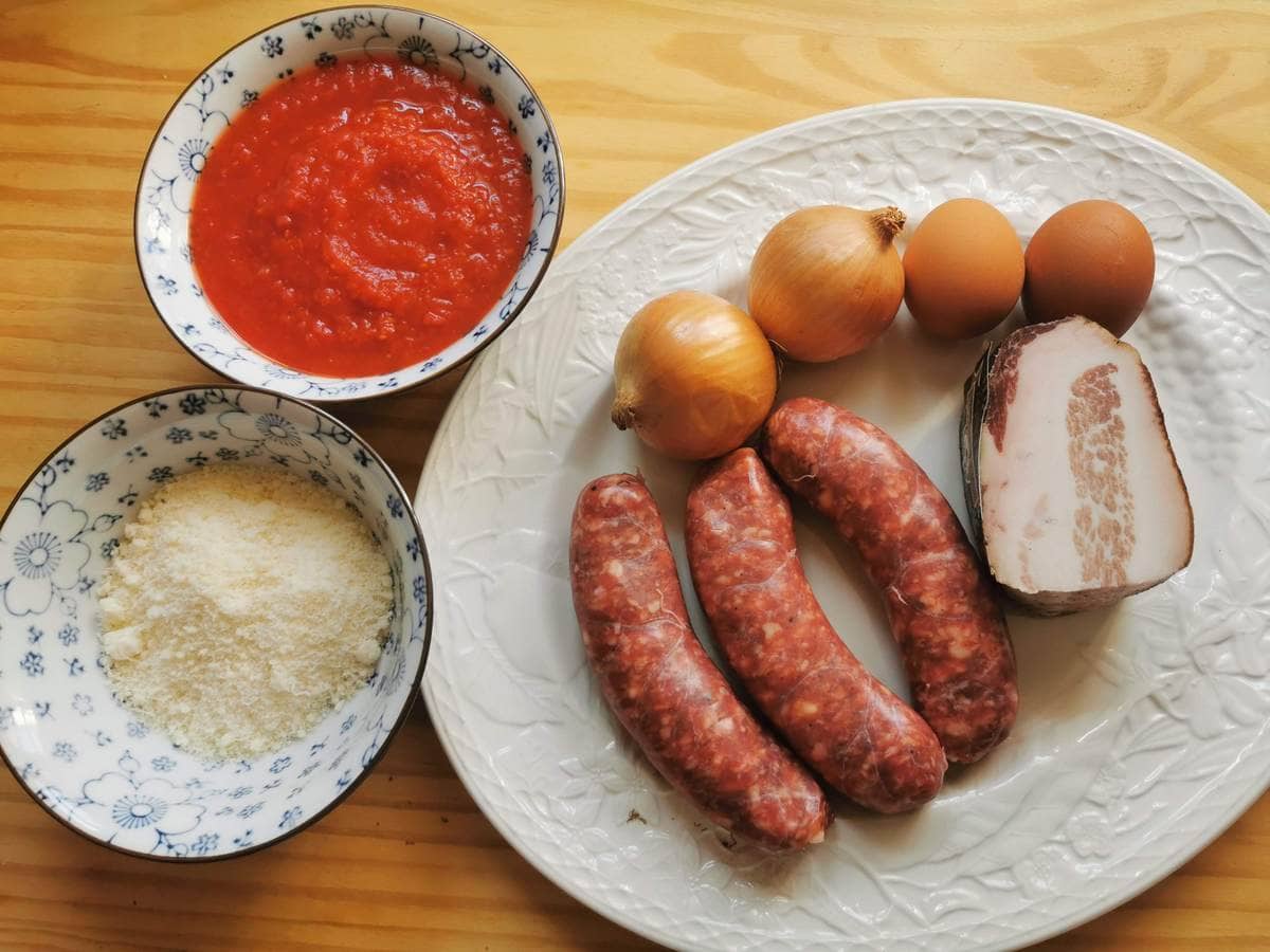 Ingredients for sausage rigatoni pasta alla zozzona on white plate; guanciale, eggs, Italian sausages, passata, onions and grated pecorino.