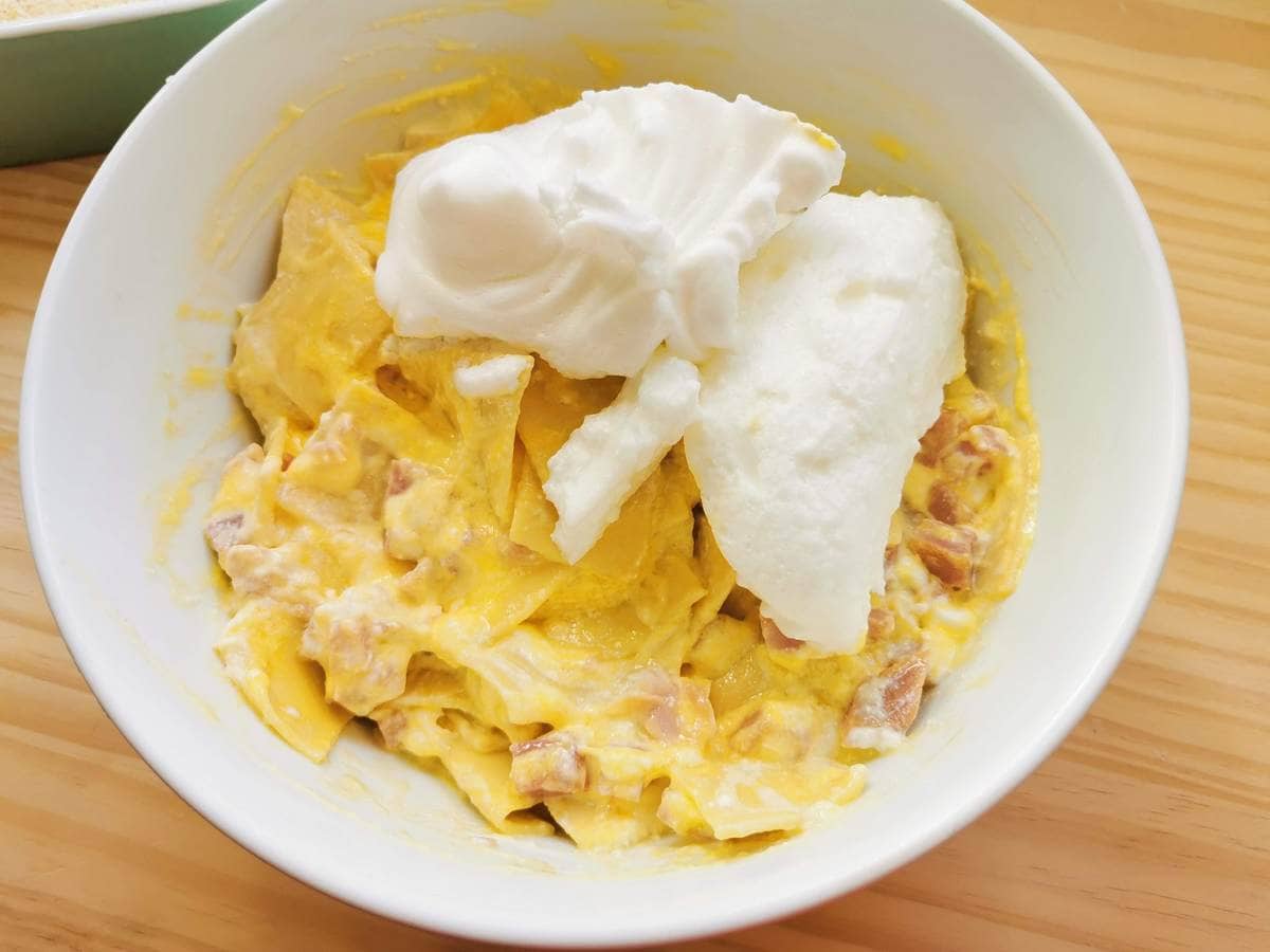 Whipped egg whites added to white bowl with other schinkenfleckerl ingredients.