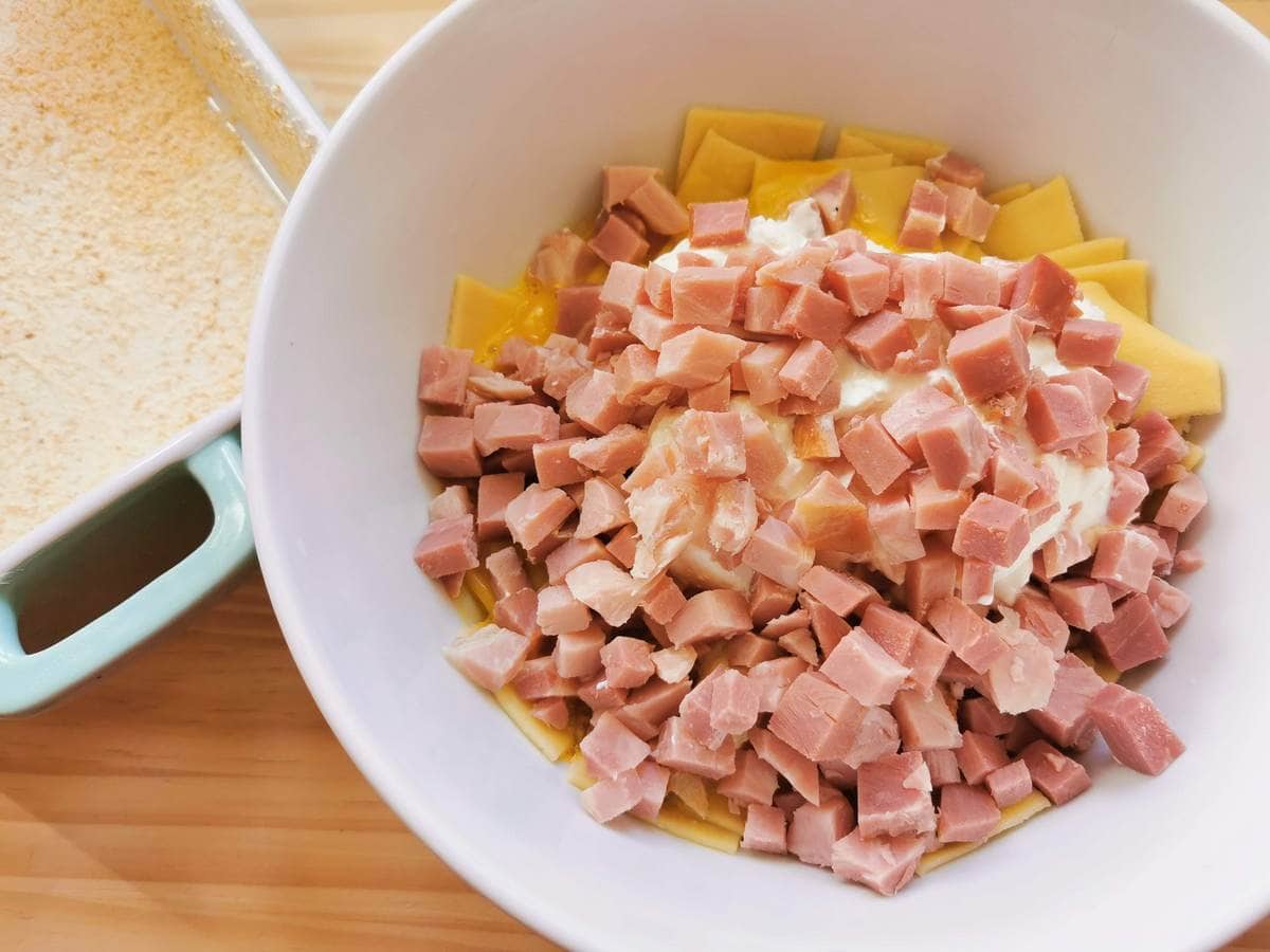 Small cubes of cooked ham added to white bowl with pasta squares, egg yolks and sour cream.