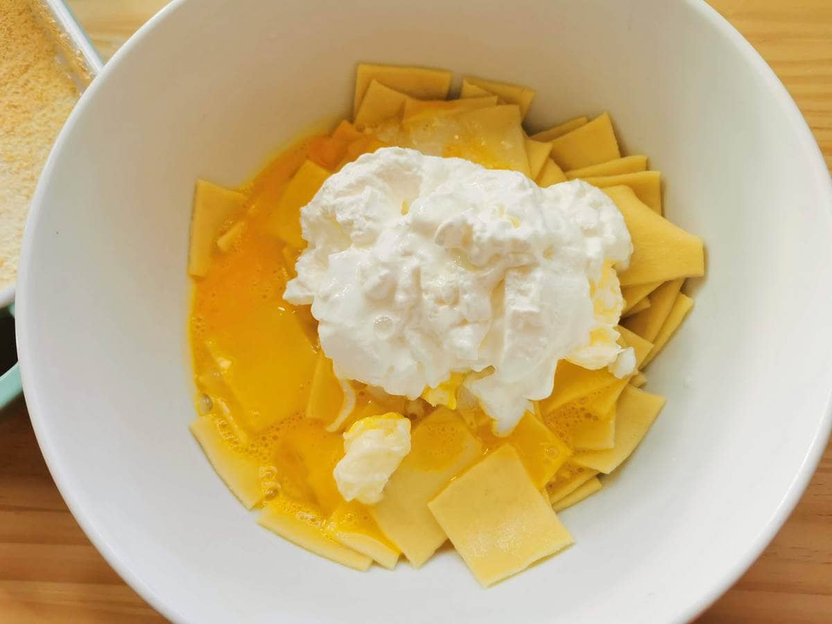 Sour cream added to white bowl with cooked pasta squares and egg yolks.