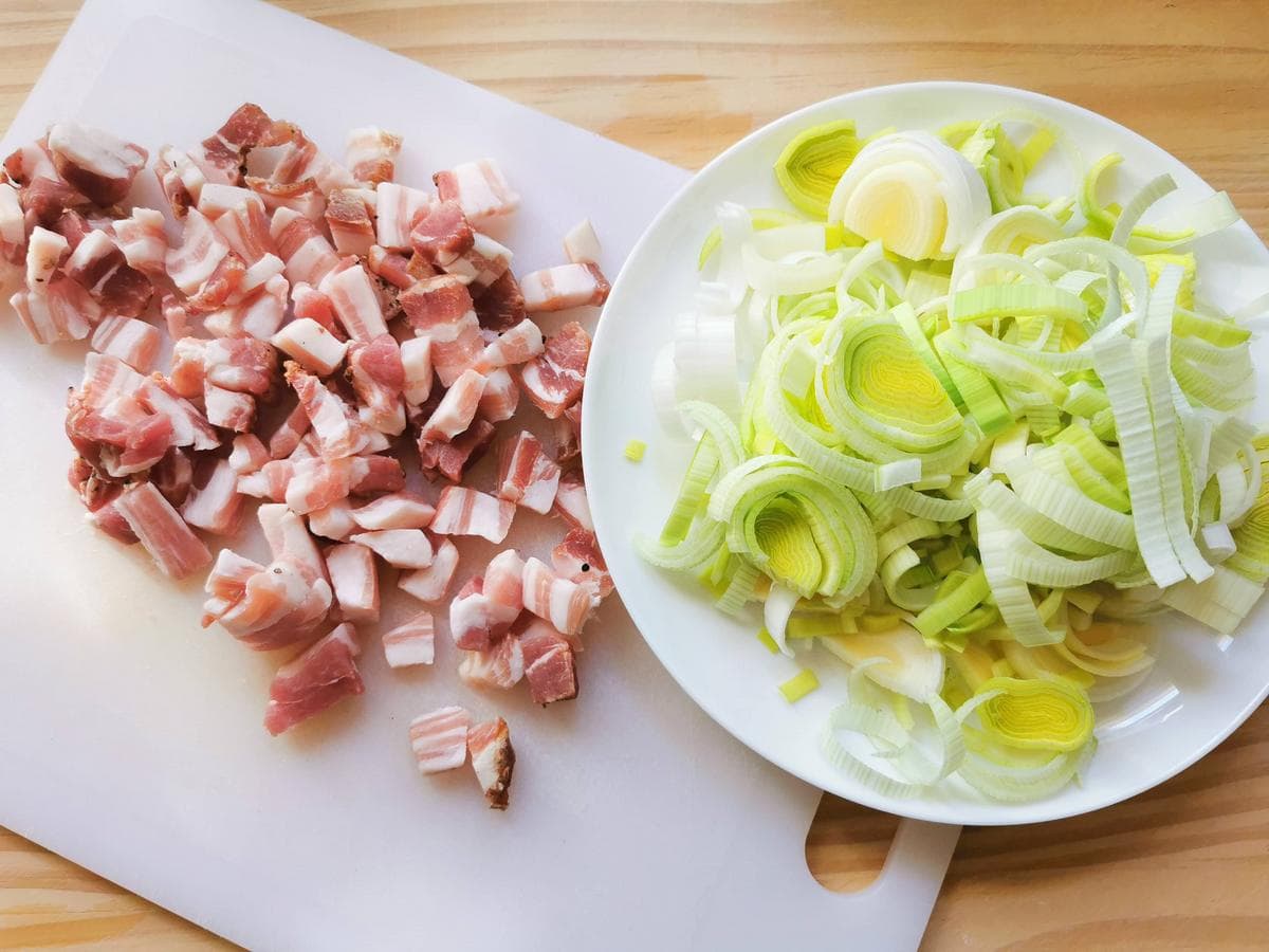 Sliced leeks on white plate and cubed pancetta on white cutting board.