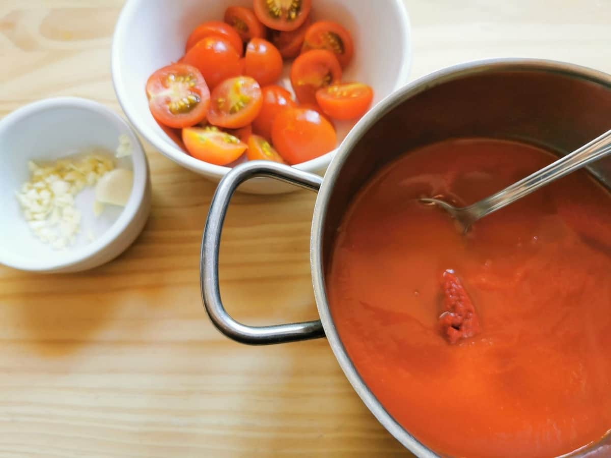 Tomato broth in saucepan. Chopped garlic and halved cherry tomatoes in white bowls.