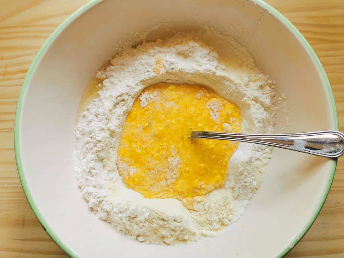Beaten eggs in the middle of flour in a bowl.