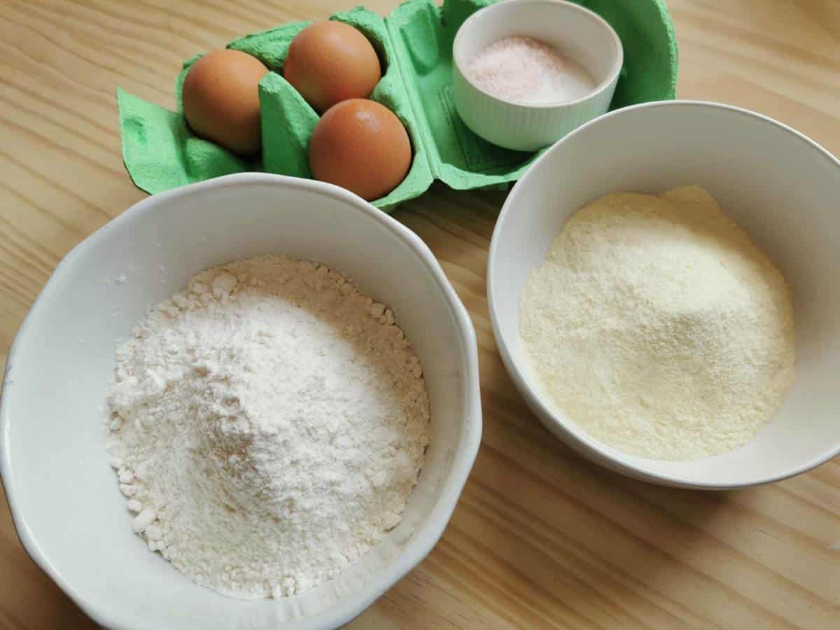 Ingredients for spaghetti alla chitarra, 2 types of flour in white bowls, eggs in a carton and salt.