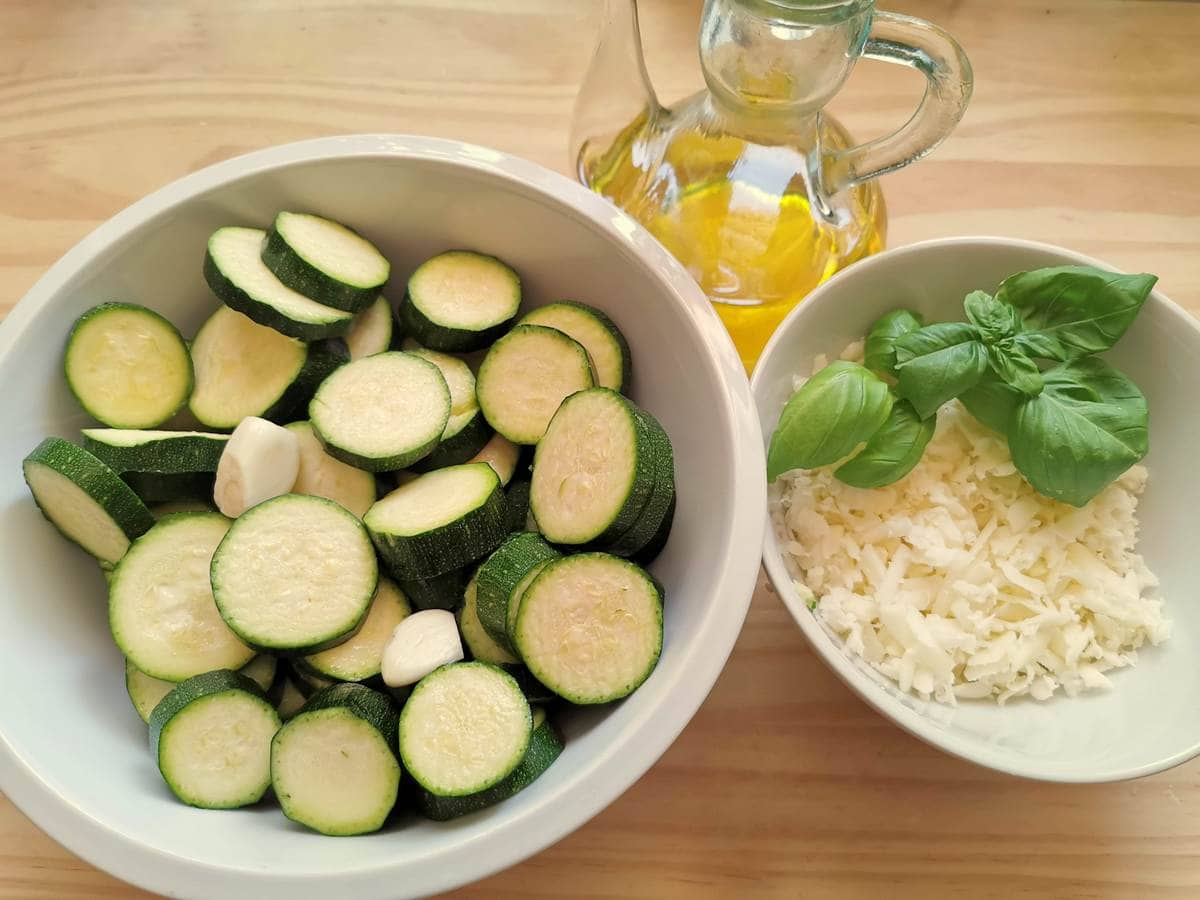 sliced zucchini and peeled garlic cloves in white bowl and grated provolone in white bowl