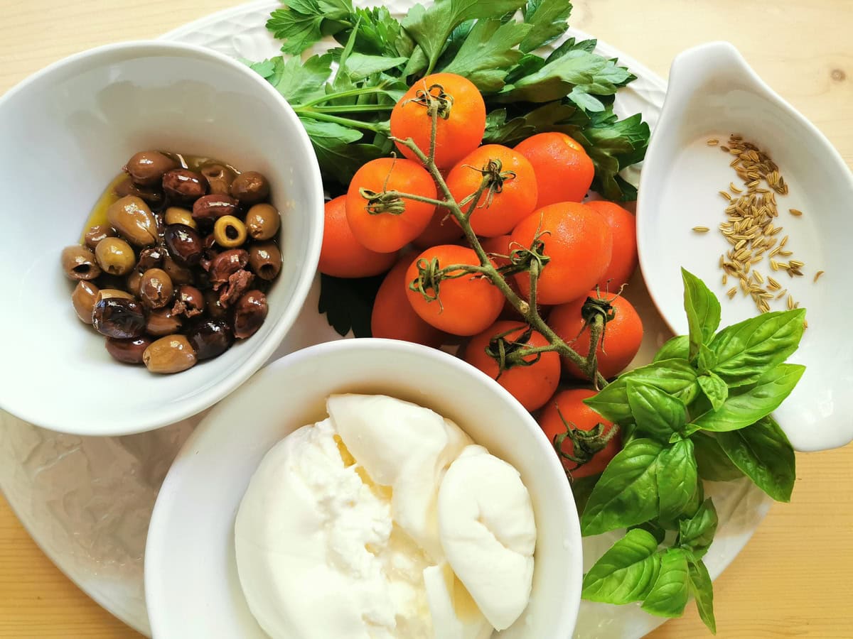 Ingredients for alla checca condiment; tomatoes, fresh basil and parsley leaves, olives, fennel seds and burrata in bowl.